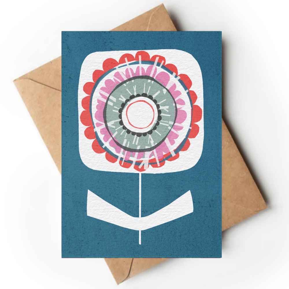 A teal art print or card with a stylized flower design, placed on top of a brown kraft envelope against a white background. The print has a dark teal-blue background with a flower motif in white, pink, red, and gray.