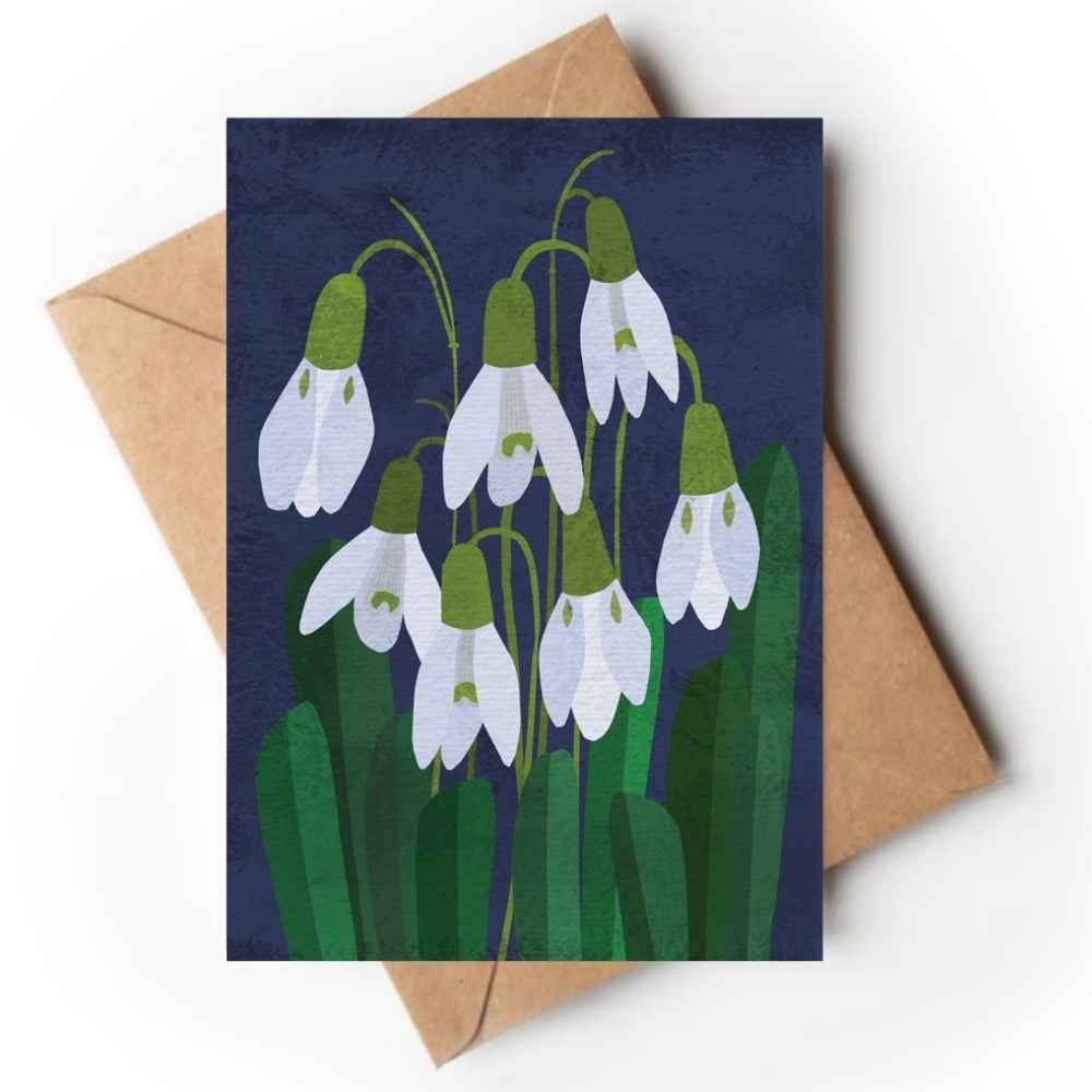 A greeting card featuring a stylized illustration of white snowdrops with green stems and leaves against a dark navy background, shown with a brown kraft envelope on a white surface.