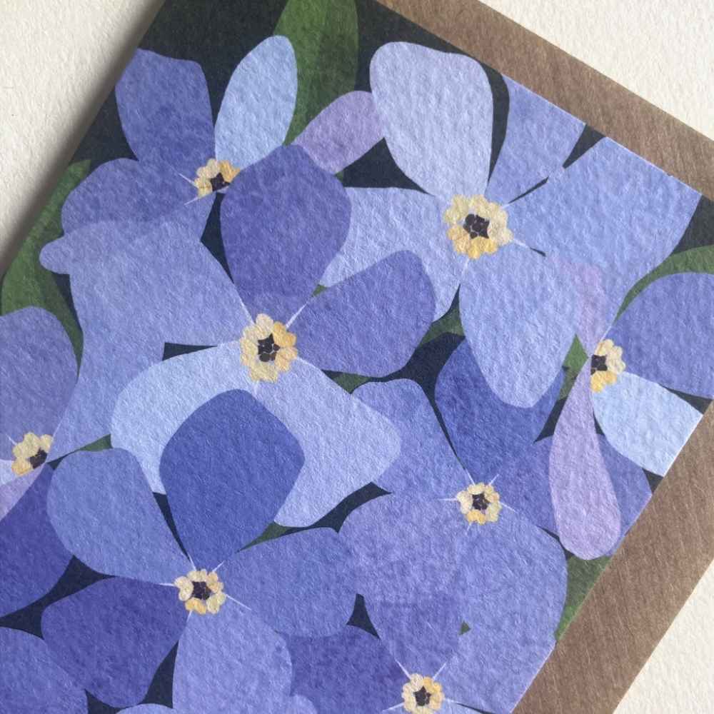 A close-up of a floral art print or card featuring layered purple and blue flowers with yellow centers and green leaves, bordered by a brown textured edge on a light neutral background.